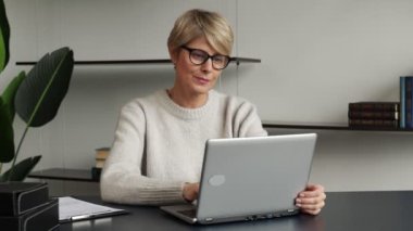 Çekici, orta yaşlı bir iş kadını dizüstü bilgisayarın önünde oturuyor ve küçük işlerini evden yürütüyor..