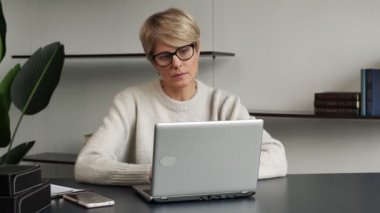 Laptop ve akıllı telefon kullanan orta yaşlı bir kadının portresi. Bir kadın ofiste çalışırken mesajlaşır.