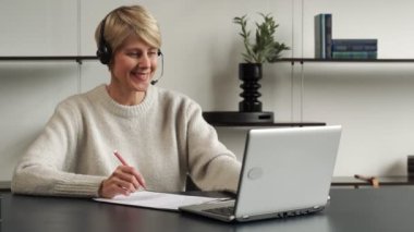 Gülümseyen orta yaşlı modern kadın kulaklık takıyor, uzaktan eğitim alıyor ya da dizüstü bilgisayarda eğitim görüyor..