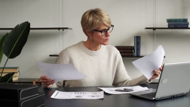 Una mujer de mediana edad sentada en un escritorio de una oficina, trabajando con una computadora portátil, está abrumada y arroja documentos sobre la mesa — Vídeos de Stock