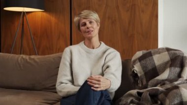 Oturma odasındaki kanepede oturan ve kameraya bakan orta yaşlı bir kadının portresi. Mutlu bir bayan internette hoş bir sohbetten hoşlanır, arkadaşlarıyla internet üzerinden iletişim kurar.