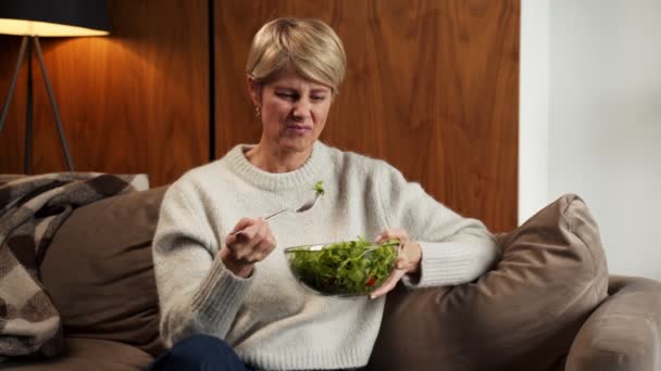 Sad middle-aged woman eats a tasteless green vegetable salad sitting on the couch — Stock Video