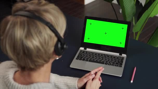 Vista sulla spalla di una donna di mezza età in cuffia, che lavora o studia su un computer portatile con schermo verde. — Video Stock