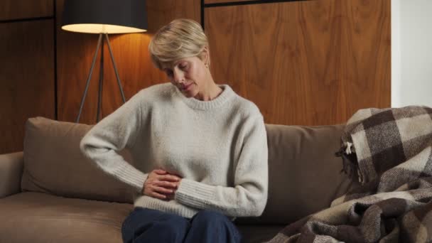Terrible abdominal pain. A sick middle-aged woman suffers from abdominal pain, holding her stomach at home while sitting on the couch — Stock Video