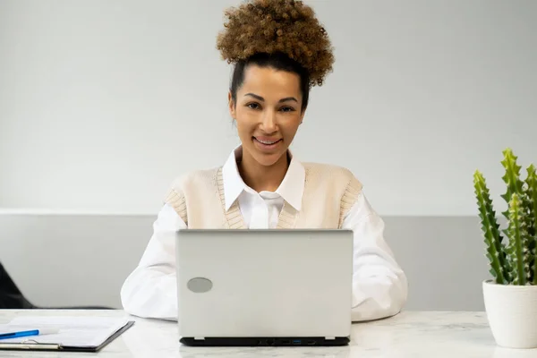Joven empresaria afroamericana utiliza una computadora portátil en el lugar de trabajo y mira a la cámara — Foto de Stock