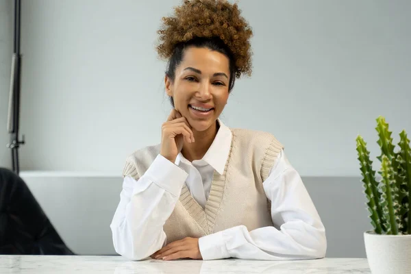 Retrato de una mujer de pelo rizado negro de moda con una sonrisa radiante sentada en un escritorio en una oficina moderna. — Foto de Stock