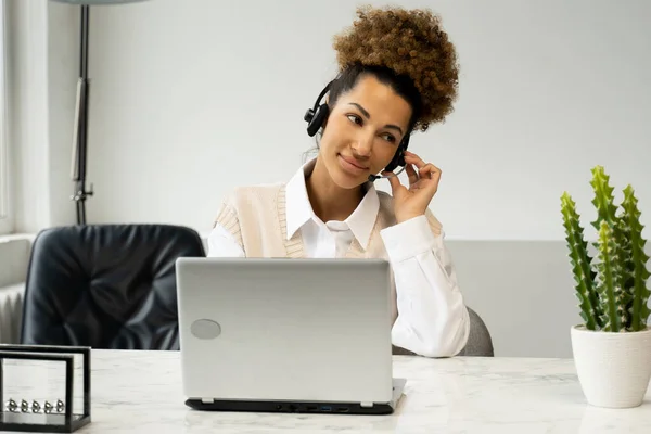 Un agente de centro de llamadas afroamericano sonriente, con auriculares con micrófono, hablando con un cliente, mirando la pantalla de un portátil. — Foto de Stock