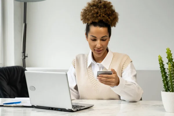 Mujer afroamericana sosteniendo una tarjeta de crédito, sentada en una mesa con un portátil, comprando en línea, haciendo pagos por Internet — Foto de Stock