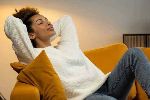 Una joven negra está sentada en un cómodo sofá en la sala de estar con los ojos cerrados y las manos detrás de la cabeza. — Foto de Stock
