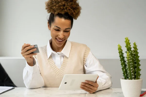 Una mujer negra sosteniendo una tarjeta de crédito hace un pago en línea usando una tableta. Mujer de pelo rizado joven sentada en un escritorio — Foto de Stock