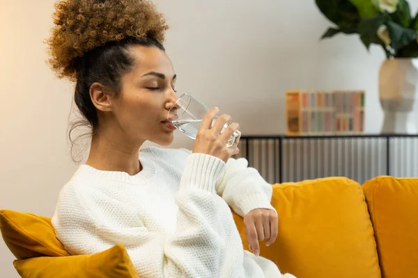 Joven mujer negra bebiendo un vaso de agua sentada en el sofá de la sala — Foto de Stock