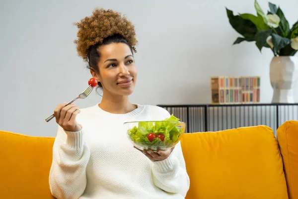 Привлекательная молодая женщина ест овощной салат из тарелки, сидя на диване дома — стоковое фото