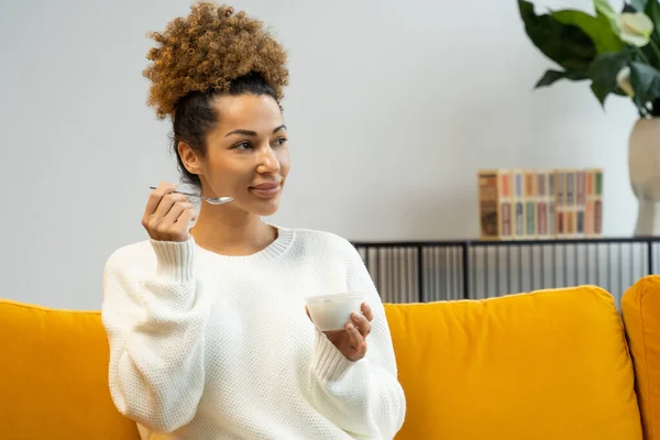 Hermosa mujer negra comiendo yogur como desayuno saludable o merienda en casa sentada en el sofá — Foto de Stock