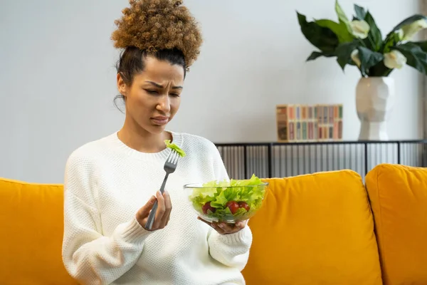 Черная женщина не любит есть овощной салат, здоровую пищу, сидя на диване — стоковое фото