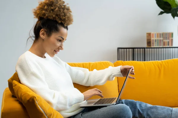 Una mujer hermosa afroamericana se sienta en un sofá amarillo y usa una computadora portátil para trabajar o busca en Internet, hace compras — Foto de Stock