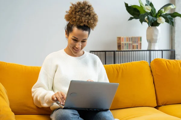 Una mujer hermosa afroamericana se sienta en un sofá amarillo y usa una computadora portátil para trabajar o busca en Internet, hace compras — Foto de Stock
