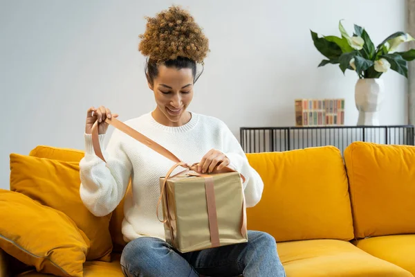 Un emocionado cliente africano joven abriendo una caja de regalo con paquetes en casa sentado en el sofá — Foto de Stock