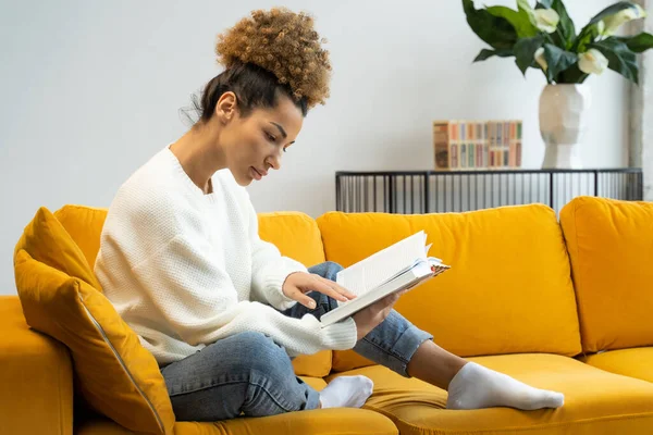 Atractiva mujer de pelo rizado negro leyendo su novela histórica favorita sentada en un acogedor sofá amarillo — Foto de Stock