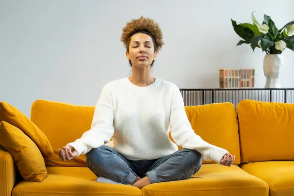 Una mujer afroamericana sentada en un cómodo sofá está haciendo yoga en posición de loto. Hermosa mujer medita en el sofá en casa — Foto de Stock