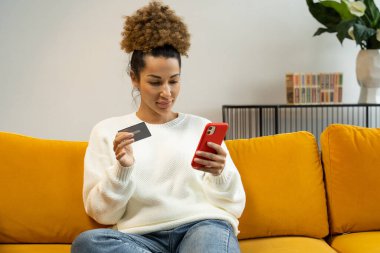 Güzel siyah bir kadın evde koltukta otururken kredi kartını internet üzerinden alışveriş ve bankacılık için kullanıyor.