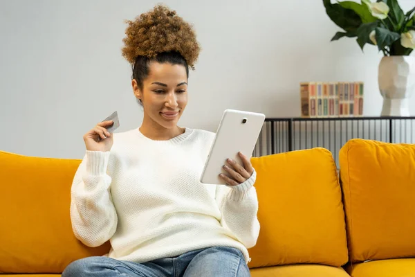 Mujer afroamericana sentada en un sofá con una tableta y una tarjeta de crédito en casa comprando online — Foto de Stock
