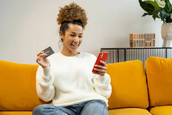 Mulher negra bonita usando um cartão de crédito para compras e bancos on-line com um telefone celular enquanto está sentado em casa no sofá — Fotografia de Stock