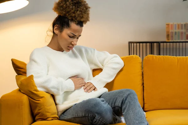 Mujer negra joven sufre de dolor abdominal en el sofá en casa. Una mujer está sentada en la cama y le duele el estómago. — Foto de Stock