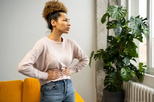 Una mujer afroamericana descontenta con el pelo rizado sufre de dolor abdominal, toca su estómago con ambas manos, mira su menstruación — Foto de Stock