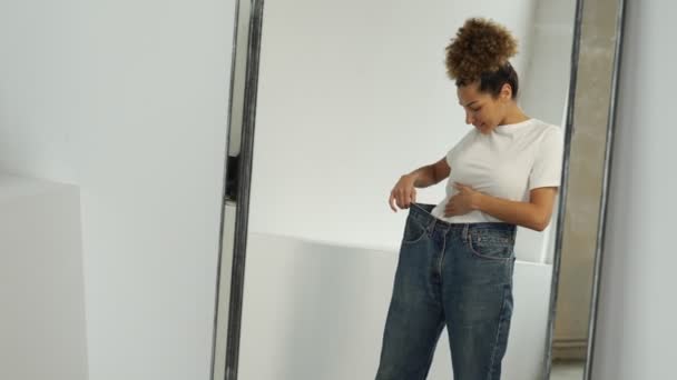 Mulher afro-americana vestida de jeans grandes antes de perder peso, olhando para a figura na frente do espelho, está muito feliz que ela perdeu peso com sucesso — Vídeo de Stock