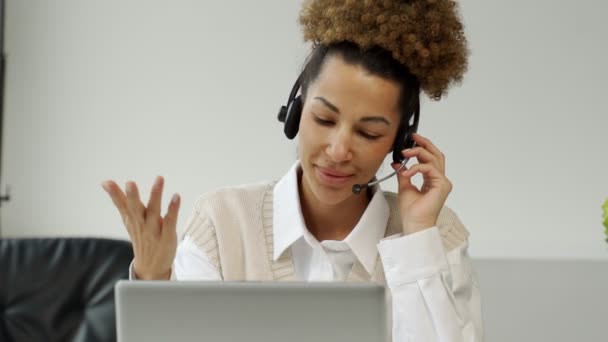 Uśmiechnięty afro-amerykański agent call center, noszący słuchawki z mikrofonem, rozmawiający z klientem, patrzący na ekran laptopa. — Wideo stockowe
