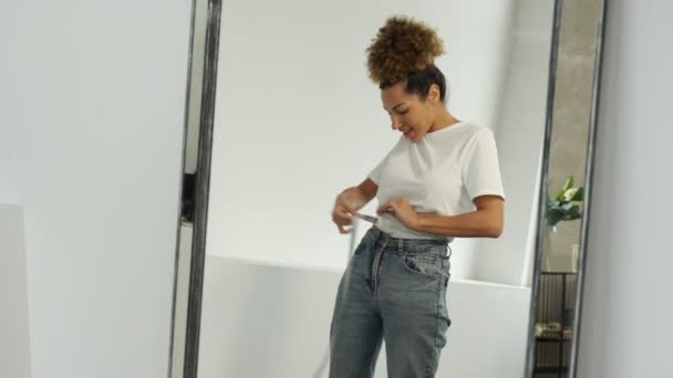 Slender black woman standing in front of a mirror measures her waist with a measuring tape after losing weight — Stock Video