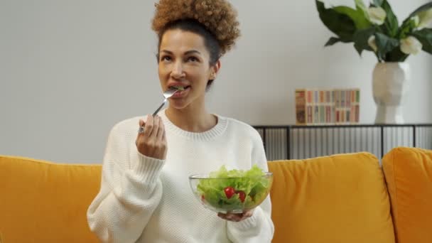 Çekici genç bir kadın evdeki kanepede oturan tabaktan sebze salatası yiyor. — Stok video