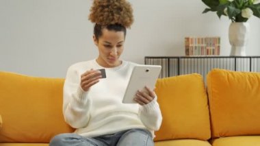 Afro-Amerikalı bir kadın elinde tablet bilgisayarla kanepede oturuyor ve internetten alışveriş yapıyor.
