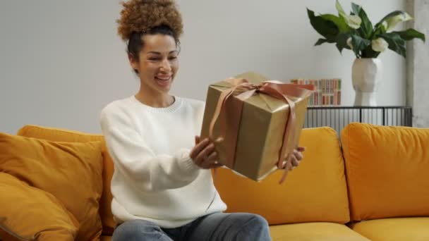 Ένας ενθουσιασμένος νεαρός Αφρικανός πελάτης ανοίγει ένα κουτί δώρου με δέματα στο σπίτι να κάθονται στον καναπέ — Αρχείο Βίντεο