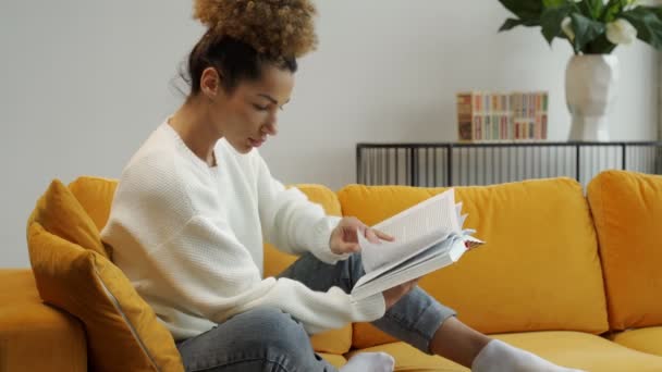 Attrayant noir bouclé femme aux cheveux lisant son roman historique préféré assis sur un canapé jaune confortable — Video