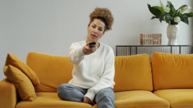 Mutlu bir Afro-Amerikan kadının portresi kanepede oturuyor ve televizyon kumandasını değiştiriyor.