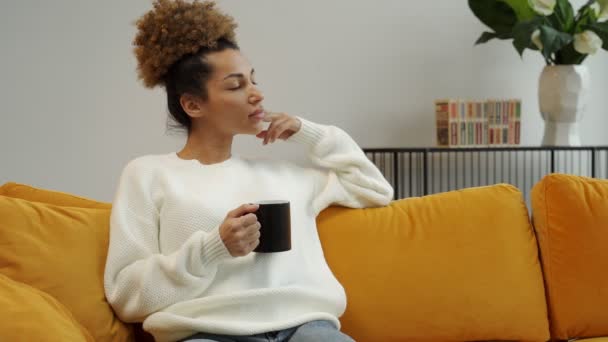 Mulher afro-americana pensativa bonita segurando uma xícara de bebida de café quente, aproveite o fim de semana em casa sentado em um sofá acolhedor — Vídeo de Stock