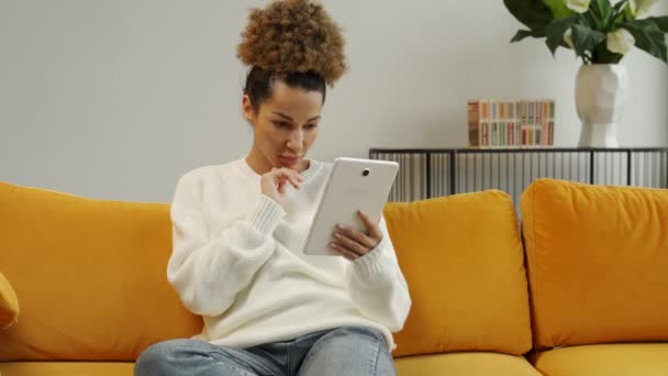 Femme afro-américaine excitée utilisant une tablette numérique, assis à la maison sur le canapé, obtient de bonnes nouvelles, gagner à la loterie, le concept de vente — Video