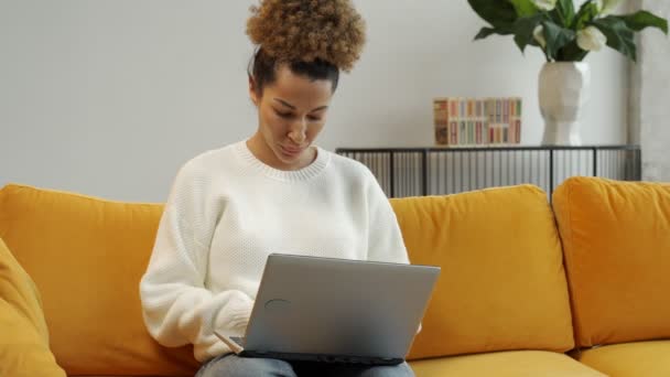 Una mujer hermosa afroamericana se sienta en un sofá amarillo y usa una computadora portátil para trabajar o busca en Internet, hace compras — Vídeo de stock