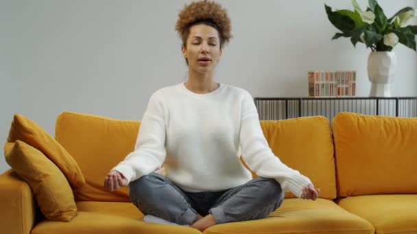 Egy afroamerikai nő, aki egy kényelmes kanapén ül, lótuszpozícióban jógázik. Gyönyörű nő meditál a kanapén otthon — Stock videók