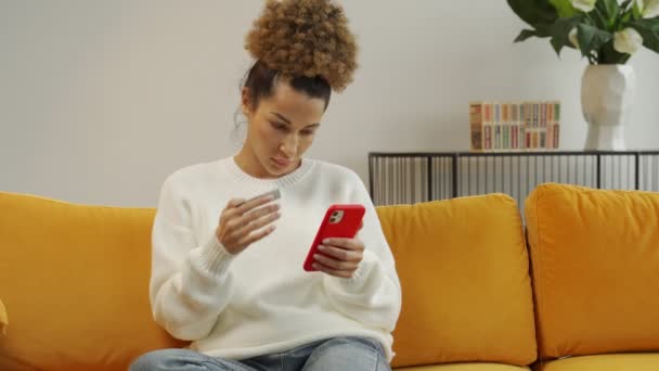 Schöne schwarze Frau mit Kreditkarte für Einkäufe und Online-Banking mit dem Handy, während sie zu Hause auf der Couch sitzt — Stockvideo