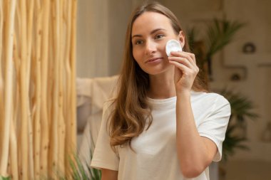 Genç ve güzel bir kadın banyoda pamukla yüzünü temizliyor. Beyaz tişörtlü bir kadın aynaya bakıyor, gülüyor ve eğleniyor.