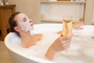 Rahatlamış bir kadın, küvette köpük banyosunun keyfini çıkarırken bir bardaktan şampanya içer.