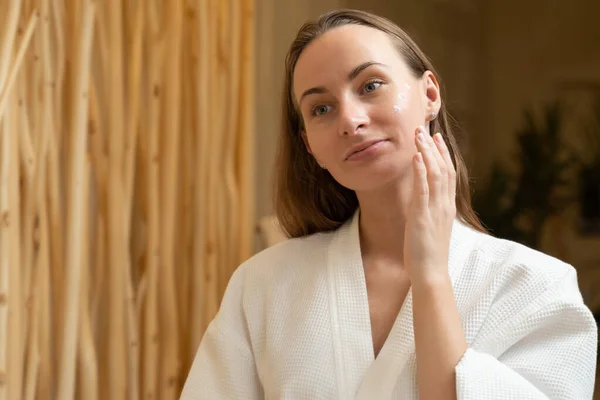 Усміхнена красива жінка в халаті кладе крем для догляду за шкірою на обличчя і дивиться у дзеркало ванної кімнати — стокове фото