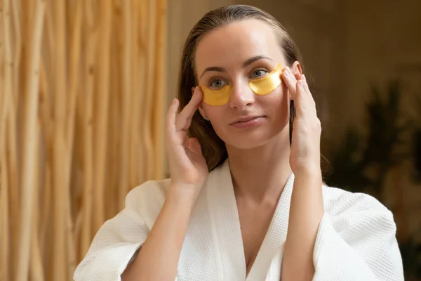 Atraktivní žena aplikuje anti-únavová maska pod jejíma očima při pohledu do zrcadla v koupelně. — Stock fotografie