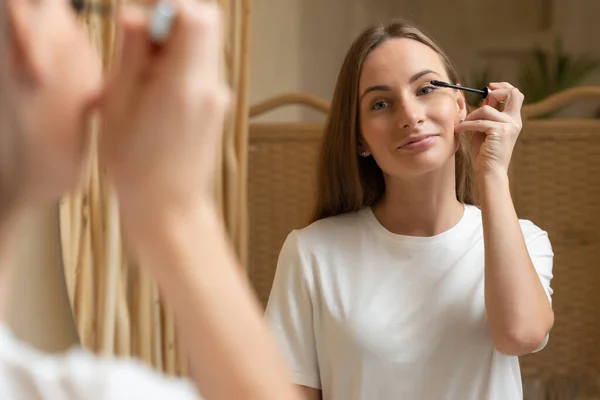 Mujer que usa rímel en la pestaña en el baño por la mañana. Mujer joven aplicando maquillaje de ojos y mirando el espejo. — Foto de Stock