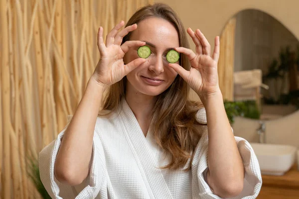 Attractive young woman in a bathrobe holding cucumber slices over her eyes while standing in the bathroom — 图库照片