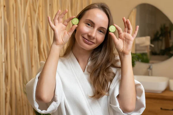 Attractive young woman in a bathrobe holding cucumber slices over her eyes while standing in the bathroom — Zdjęcie stockowe