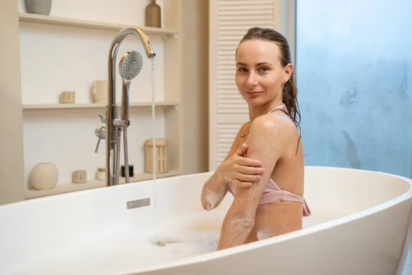 Fiatal nő alkalmazása hámlasztó bozót a vállán zuhany után, miközben ül a fürdőszobában — Stock Fotó
