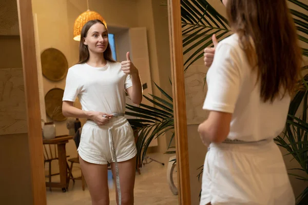 İnce bir kadın aynanın önünde duruyor ve kilo verdikten sonra belini ölçüyor.